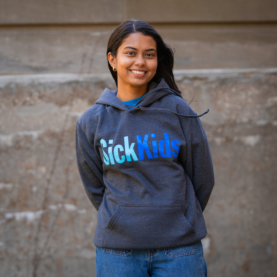 An adult female wearing a dark grey pullover hoodie with 'SickKids' text in blue on the front, standing against a stone background.