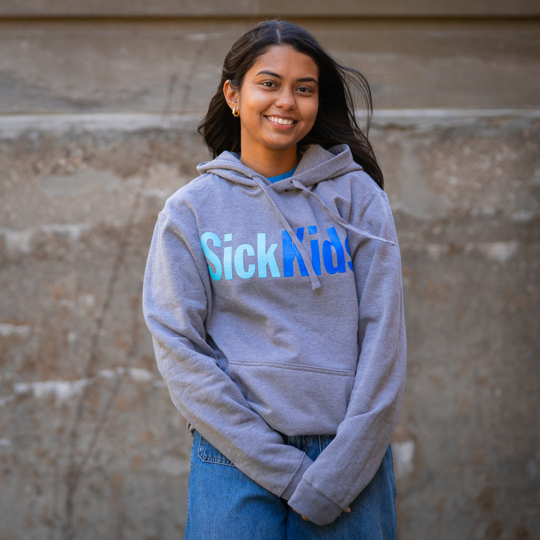 An adult female smiling and wearing a light grey pullover hoodie with 'SickKids' text in blue on the front.