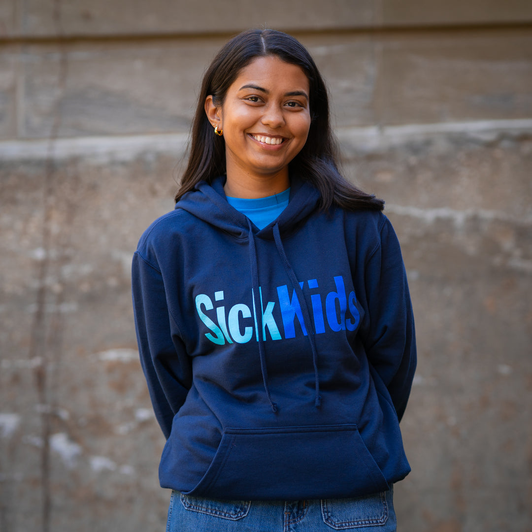 An adult female smiling and wearing a navy blue pullover hoodie with the text 'SickKids' on the front.