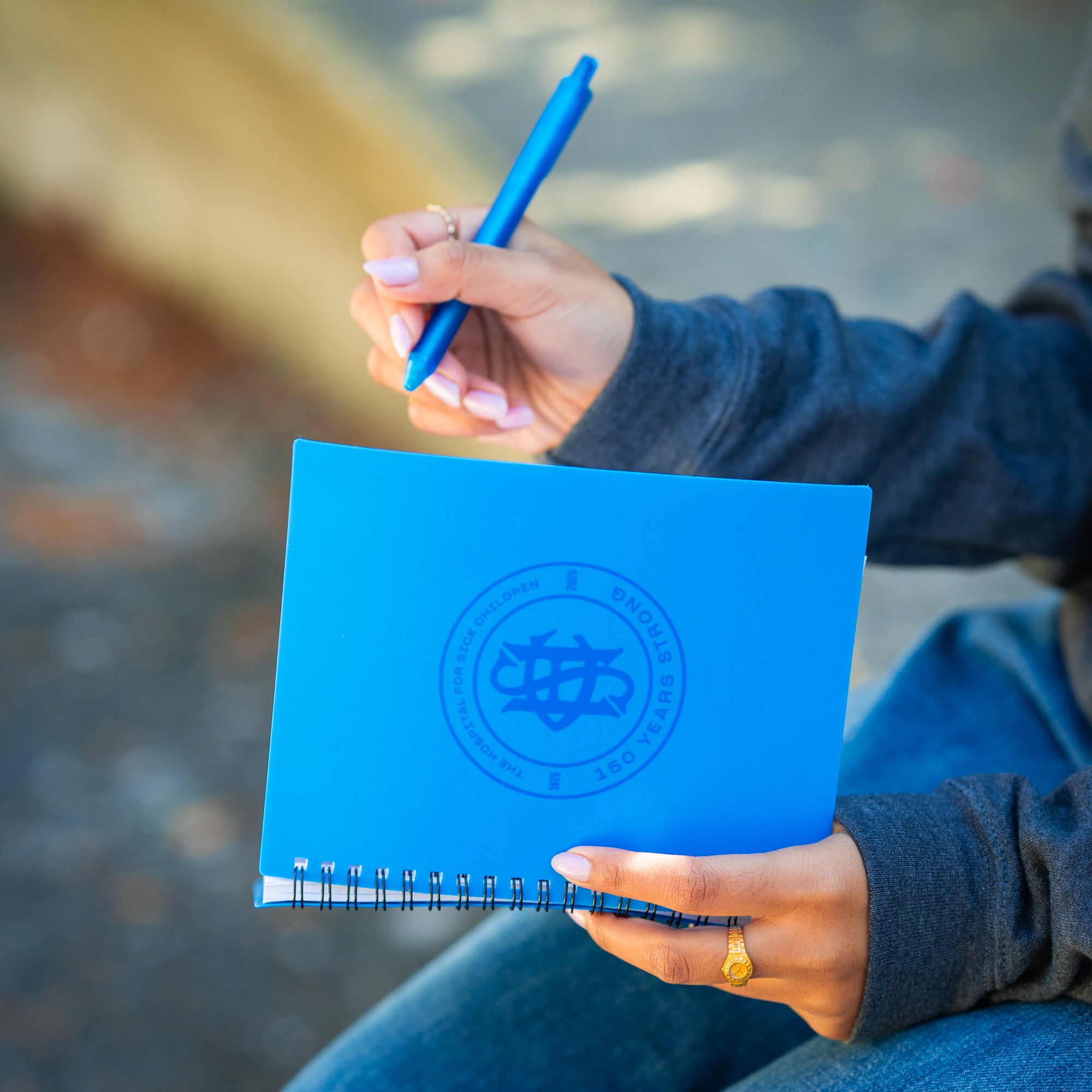A person holding a blue notebook with a circular SickKids logo and a pen, sitting outdoors.