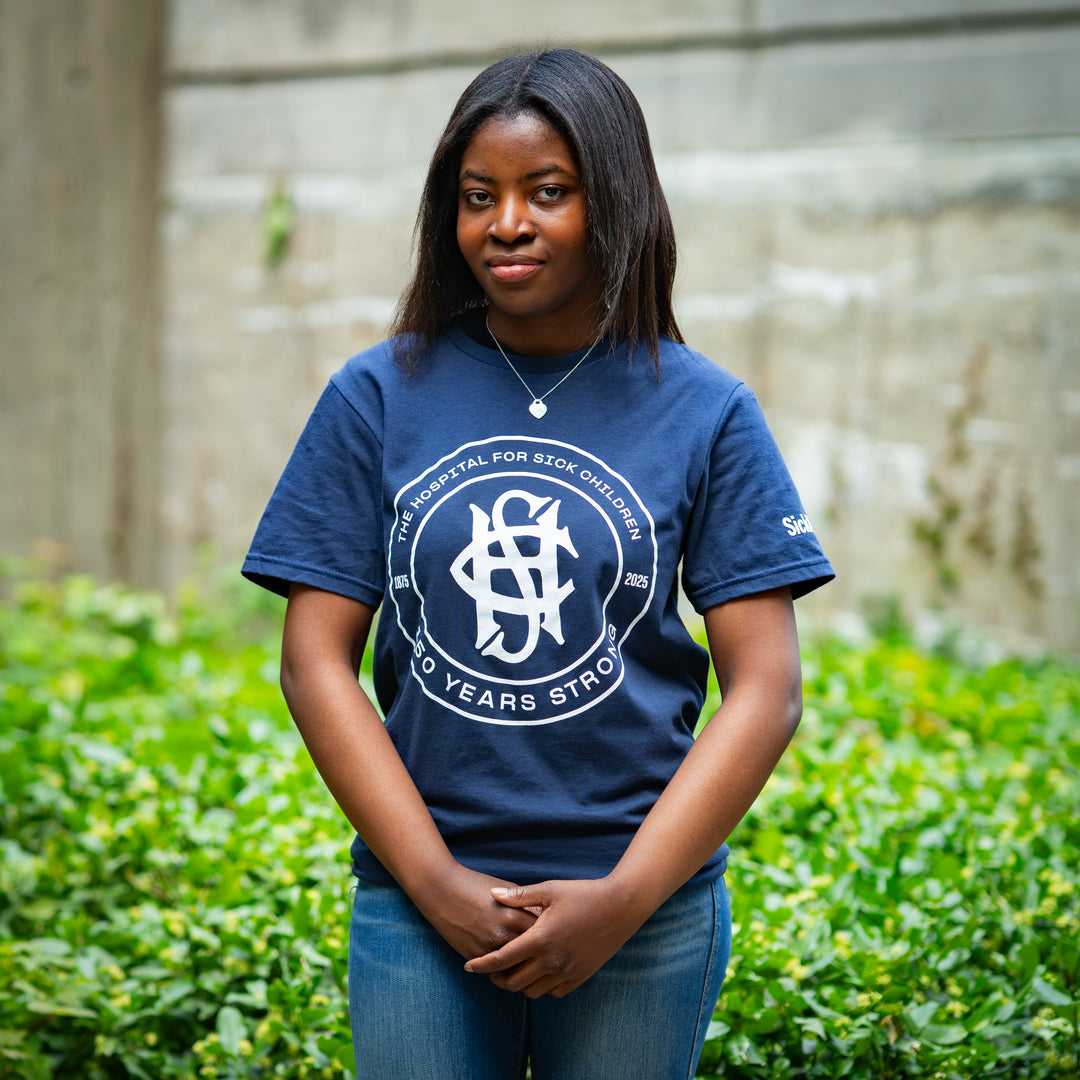 Person wearing a blue t-shirt with a circular SickKids logo and text, standing outdoors with greenery in the background