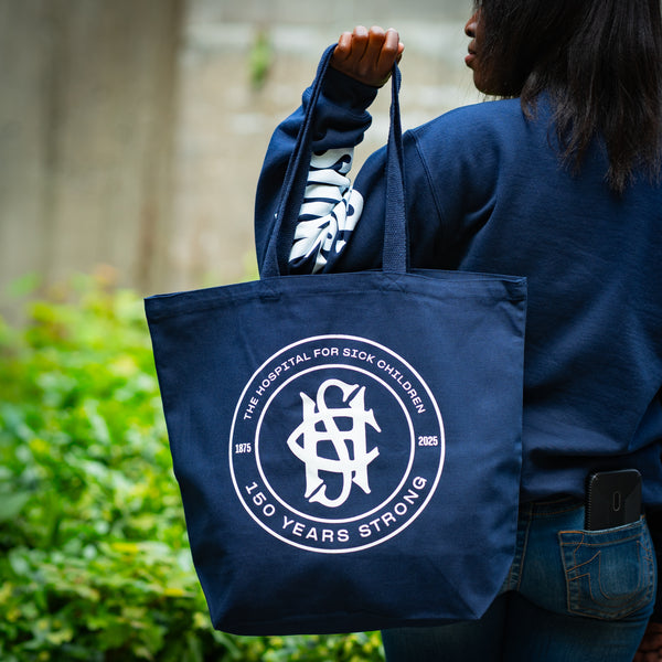 Person holding a navy blue tote bag with a logo and text, standing outdoors.