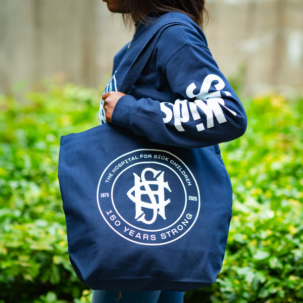 Person holding a navy blue tote bag with a logo and text, standing in front of green bushes.