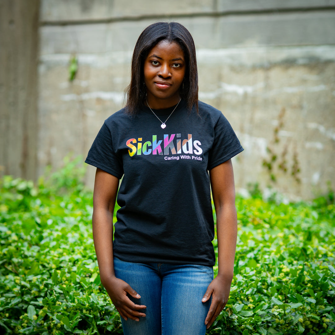 Person wearing a black 'SickKids' t-shirt standing outdoors with greenery and a concrete wall in the background.
