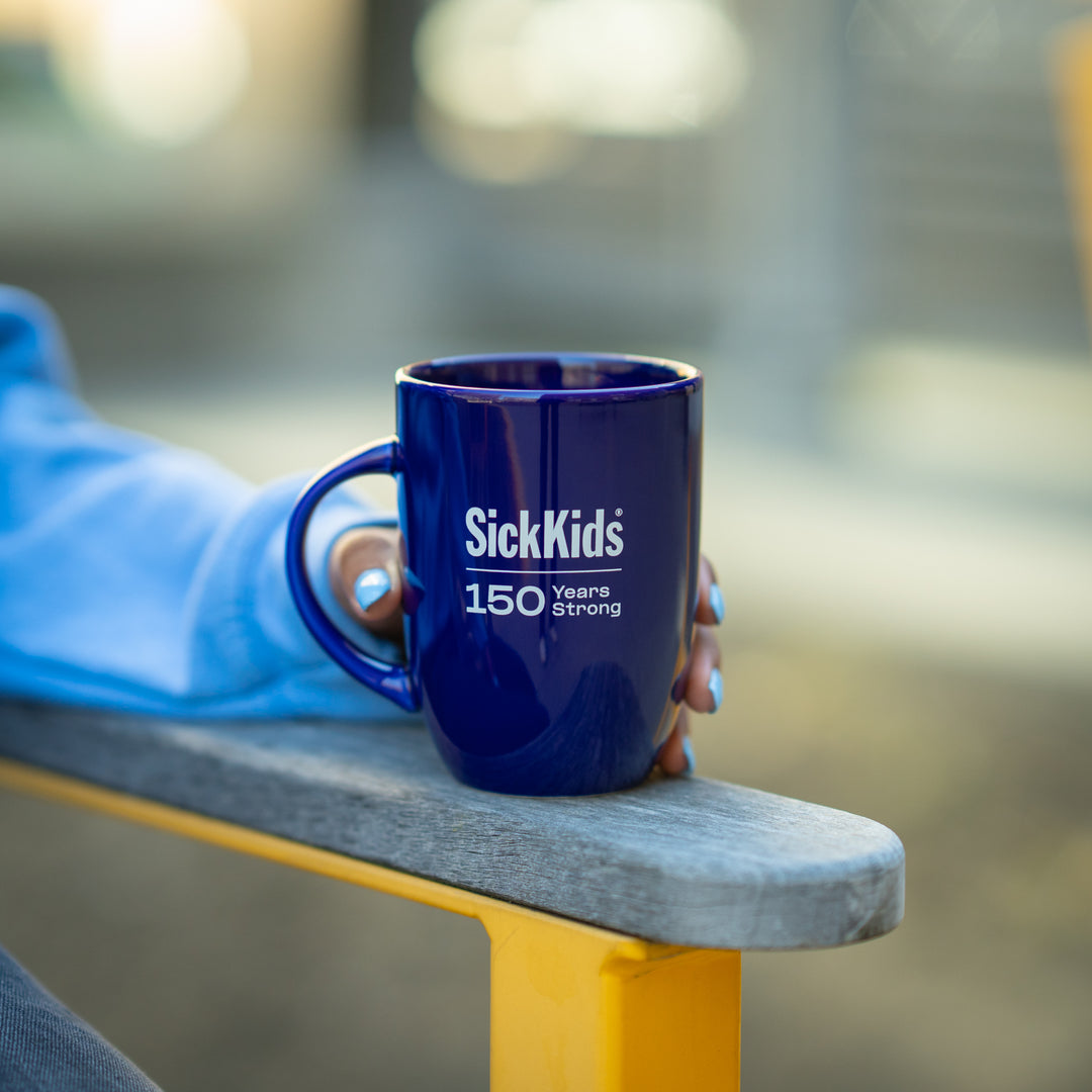 A blue ceramic mug with the SickKids 150 years strong logo printed on it, placed on a wood bench.