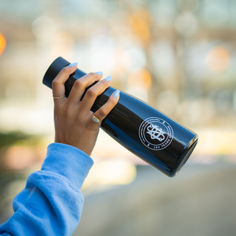 A hand holding a black, double-wall stainless steel water bottle with a premium commemorative logo, featuring a spout lid for 360-degree drinking.