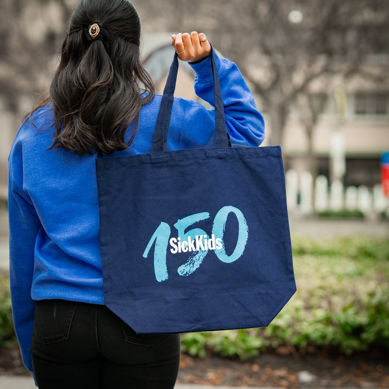 Person holding a blue tote bag with '150 SickKids' text outdoors.