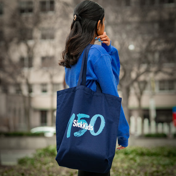 Person holding a blue tote bag with '150 SickKids' text outdoors.