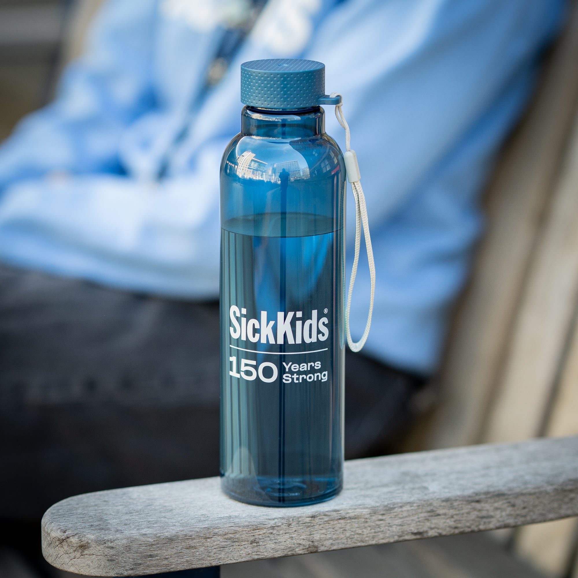 A blue water bottle with a wrist strap and the SickKids 150 years strong logo, placed on a wooden surface.