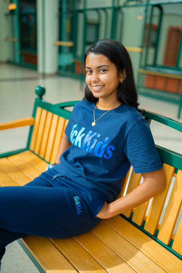 A person wearing a navy blue t-shirt with the text 'SickKids' in blue font while sitting on a bench with their hands in their pockets.