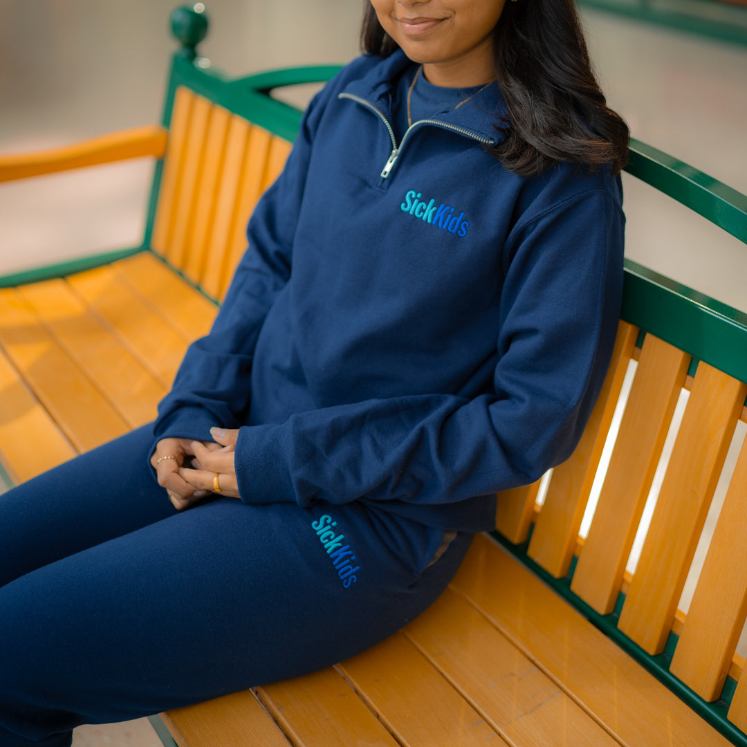 Adult wearing navy blue fleece joggers with 'SickKids' logo on the left leg, sitting on a bench.