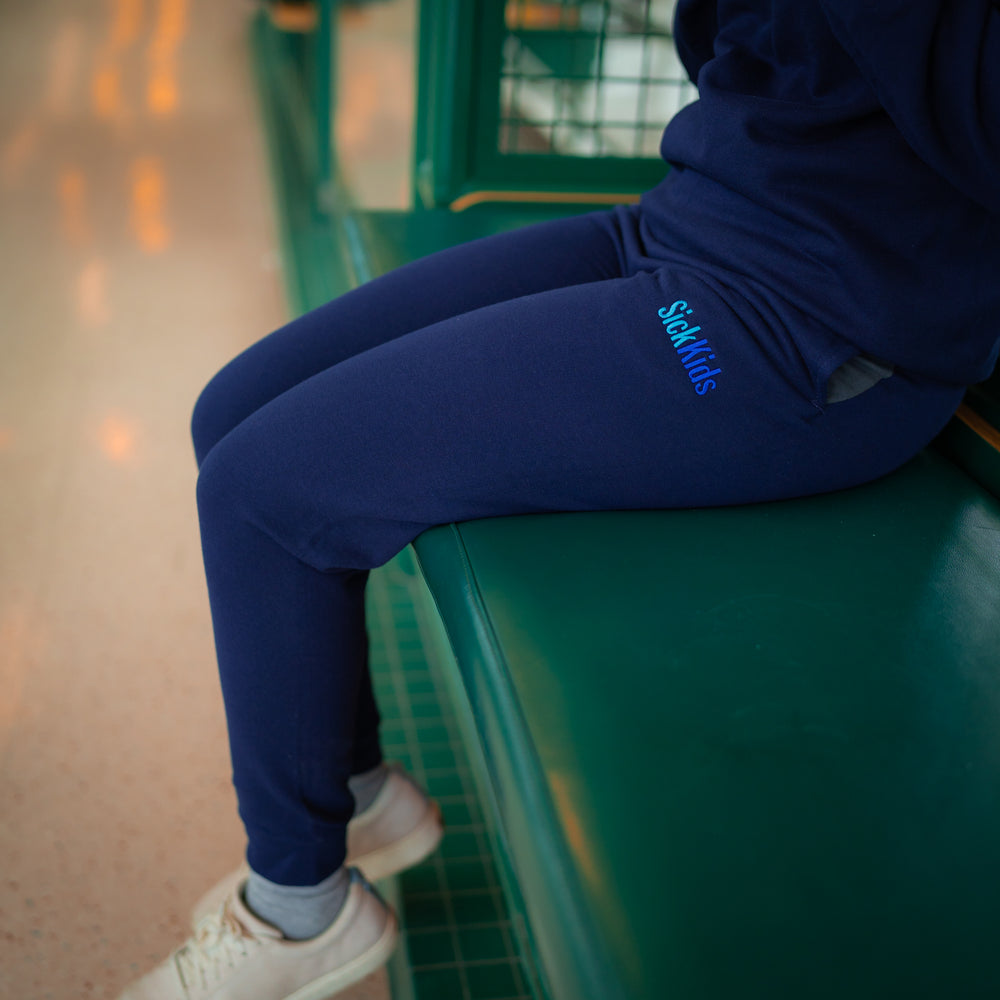 Adult wearing navy blue fleece joggers with 'SickKids' logo on the left leg, sitting on a bench.