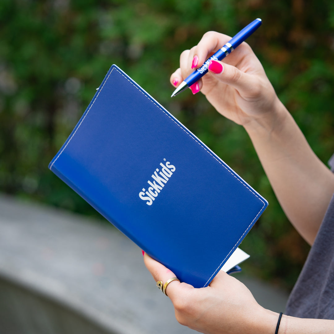 Person holding a blue notebook with 'Sick Kids' branding and a pen outdoors.