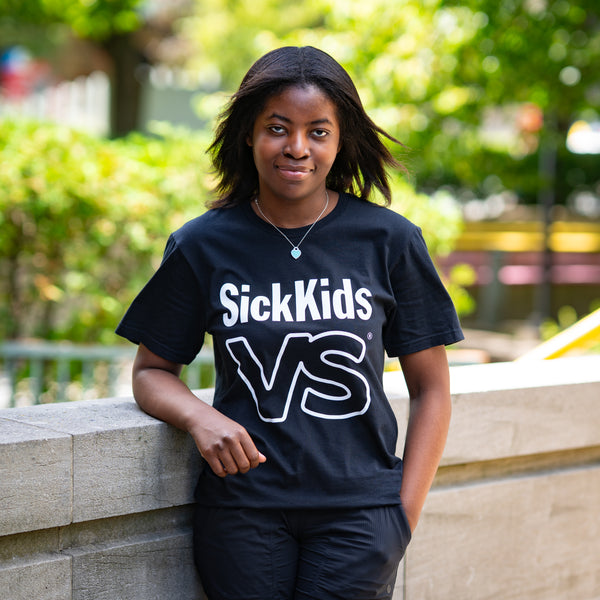 Person wearing a black 'SickKids VS' t-shirt outdoors with greenery in the background
