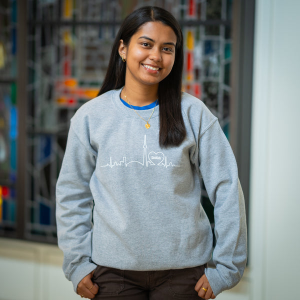A woman smiling and wearing a light grey crewneck sweater with a skyline design in the center.