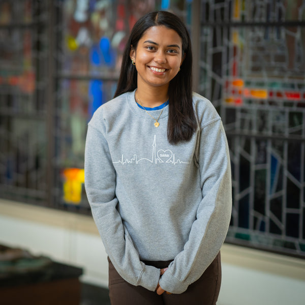A woman smiling and wearing a light grey crewneck sweater with a skyline design in the center.