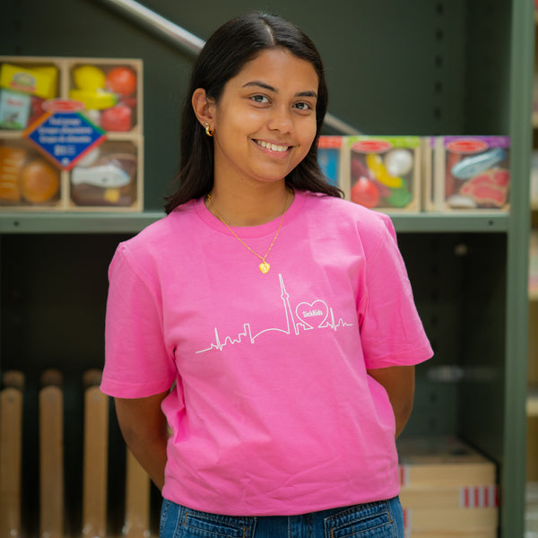 A person wearing a hot pink t-shirt with a skyline logo standing in front of a shelf full of toys.
