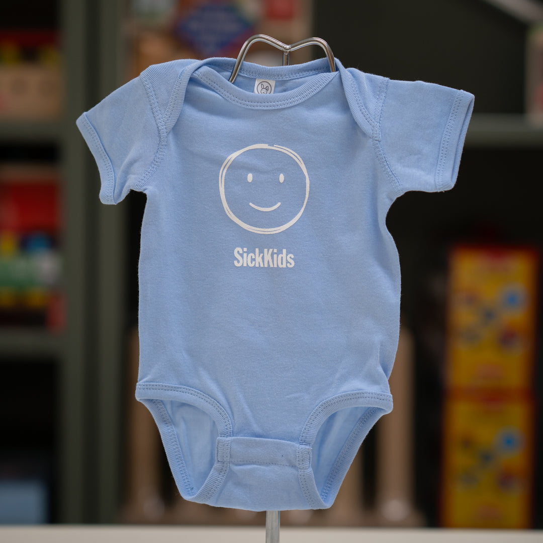 A light blue infant onesie with a smiley face and the text 'SickKids' on the front, displayed on a stand.