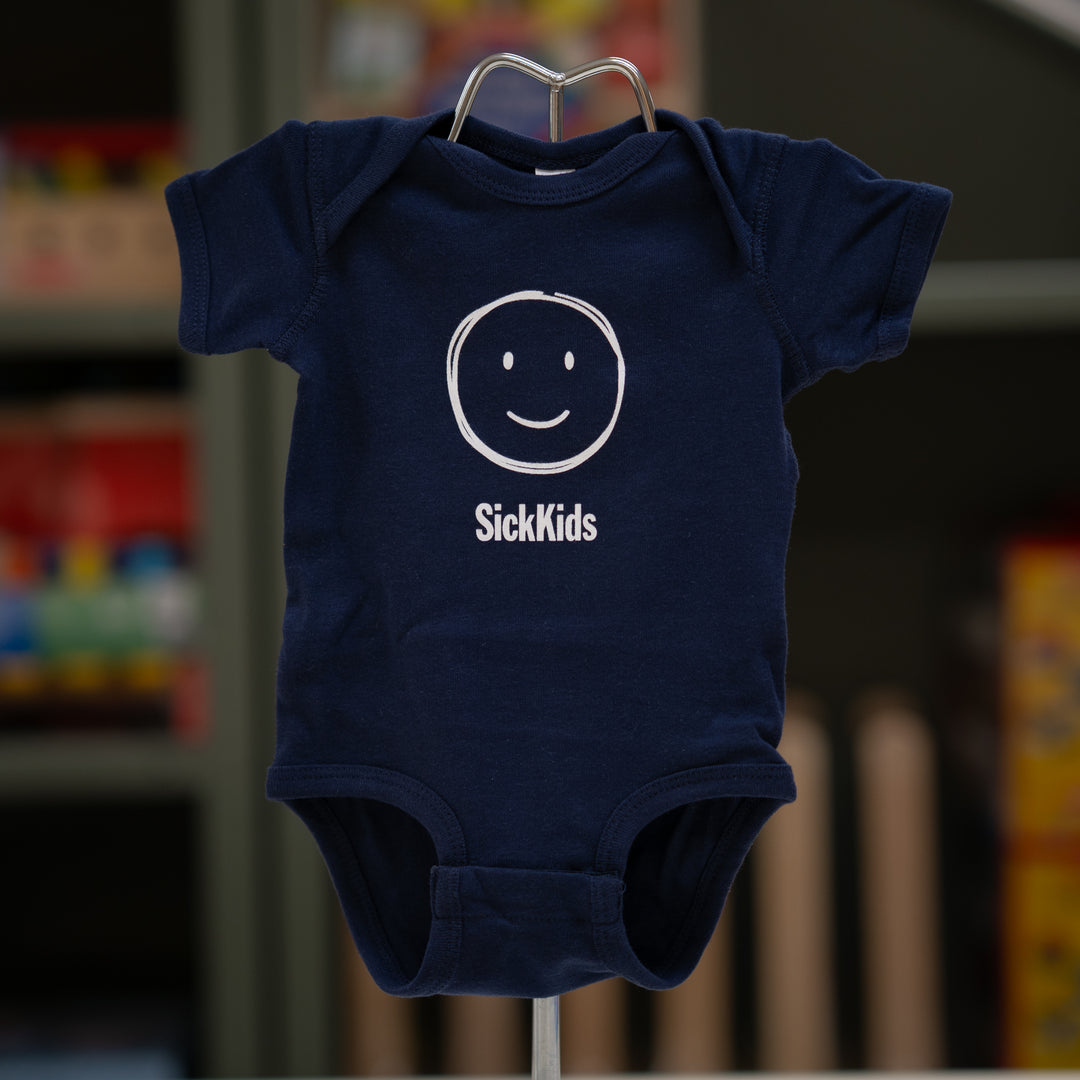 A navy blue onesie with a smiley face and SickKids logo infant of a blurry background.