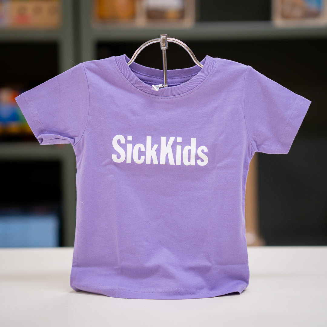 A lavender t-shirt with 'SickKids' text on a stand against a blurred background