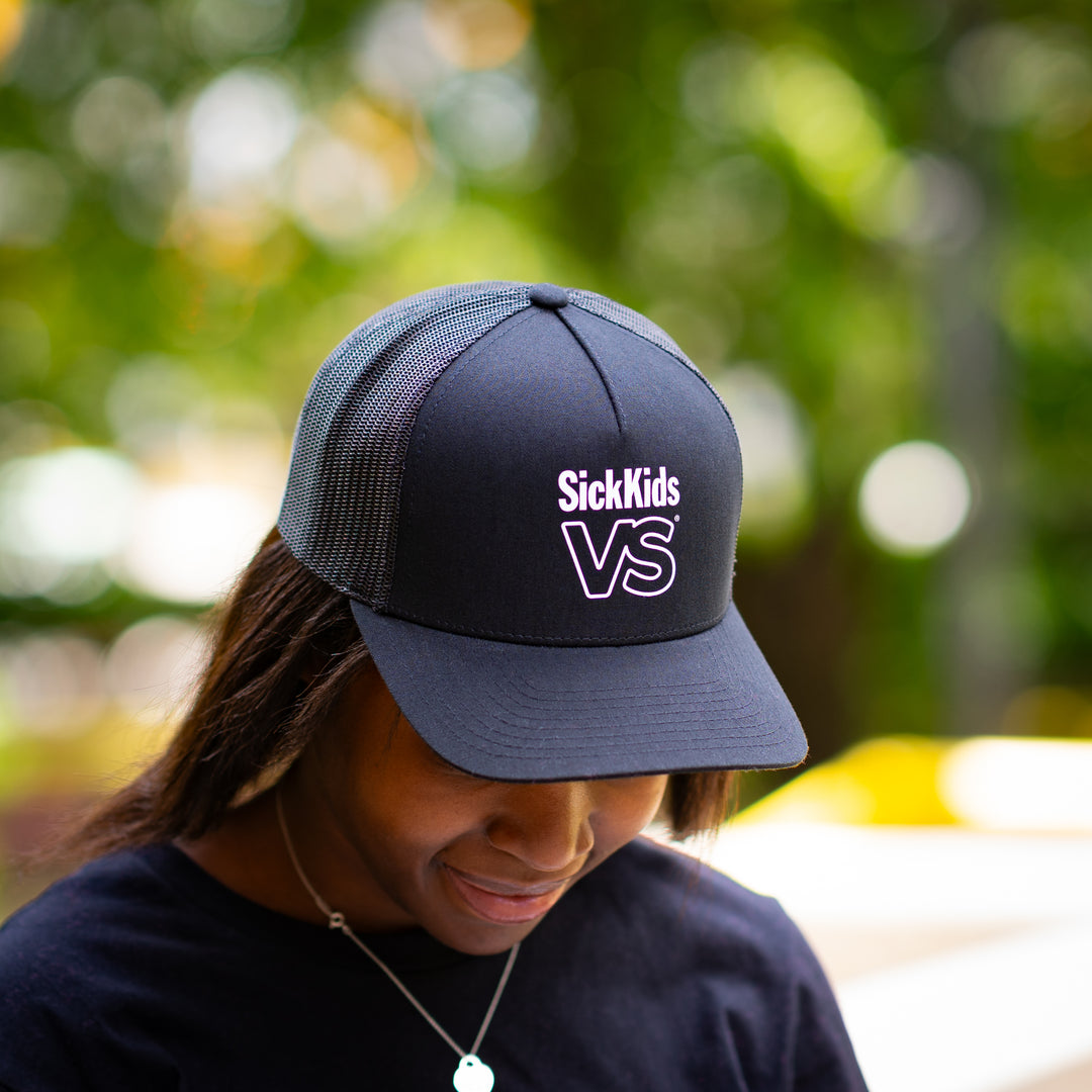 Person wearing a black baseball cap with 'SickKids VS' logo, outdoors with blurred greenery in the background.