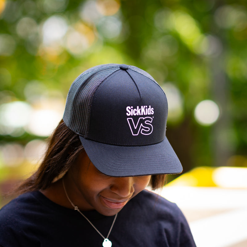 Person wearing a black baseball cap with 'SickKids VS' logo, outdoors with blurred greenery in the background.