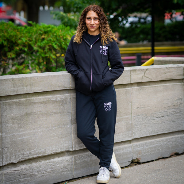 Person wearing a black zip hoodie and matching sweatpants with 'SickKids VS' logo leaning against a stone wall outdoors.