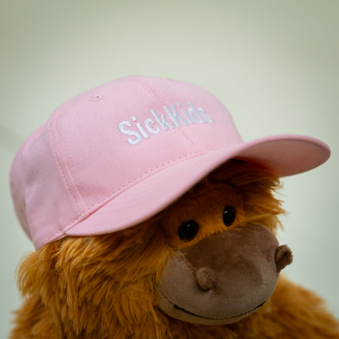 A pink baseball cap with the text 'SickKids' placed on a plush toy's head.